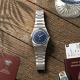 Montre automatique Belvans Daylee cadran bleu posée sur une table en bois avec accessoires de voyage.