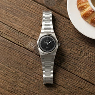 Montre automatique Belvans Daylee cadran noir posée sur une table en bois près d’un café et d’un croissant.
