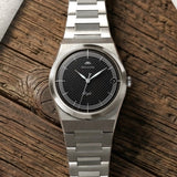 Montre Belvans Daylee cadran noir et bracelet acier posée sur bois.